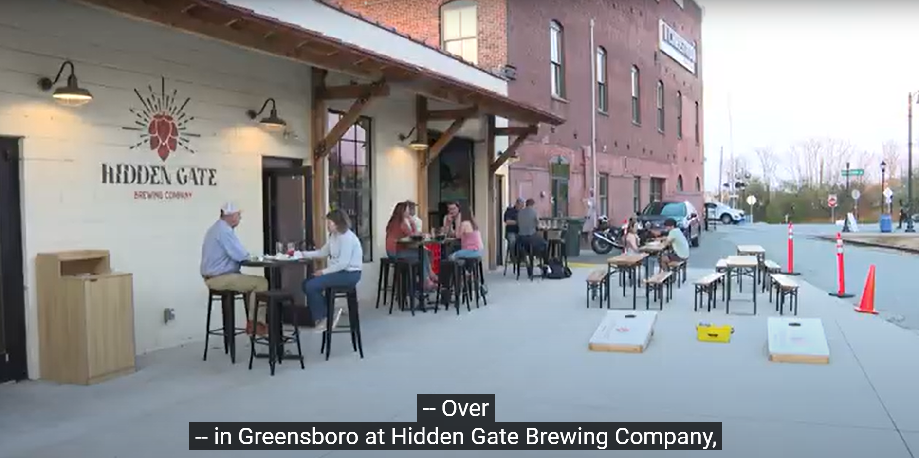 Outdoor patio area of Hidden Gate Brewing Company in Greensboro with patrons seated at high-top tables, bench seating, and cornhole boards laid out.