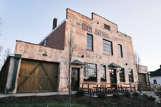 Exterior view of Oden Brewing Company’s two-story historic brick building featuring wooden garage doors, industrial windows, and outdoor patio benches.
